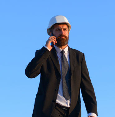 Engineers and architects in formal suits, holding black and brown briefcases, wearing white and orange helmets, walking confidently under a blue sky. One person talks on the phone, embodying a professional business and construction concept